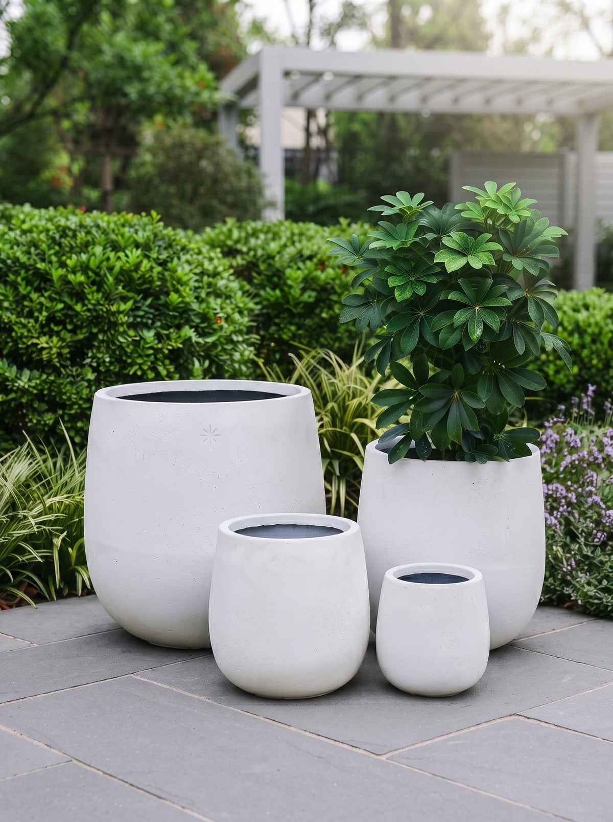 Three matte white planters in different sizes arranged on a stone patio in a landscaped garden with green shrubs and a pergola in the background.